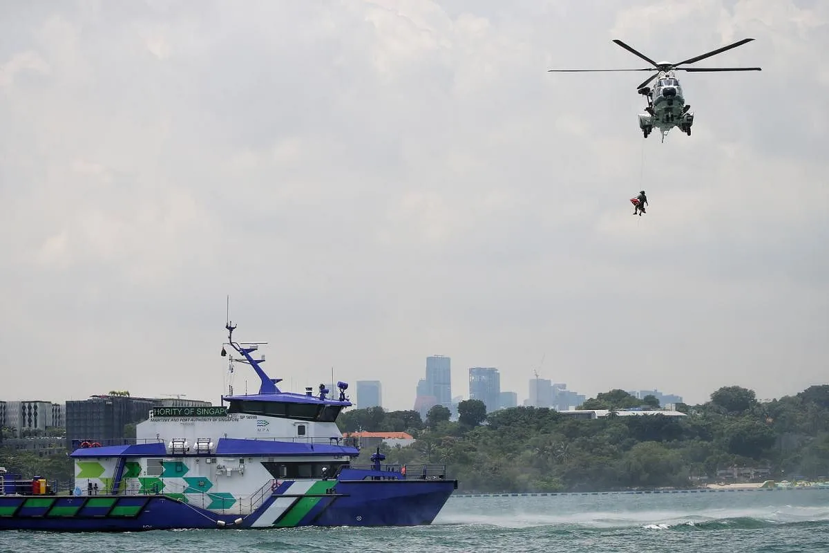 A helicopter hovers over water, hoisting a person. Below is the MPA Guardian with the city skyline behind.